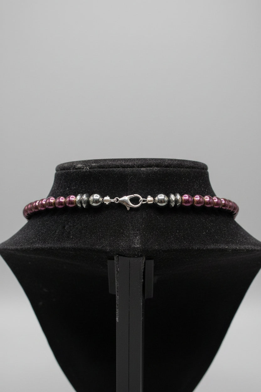 A close-up of a beaded bracelet or necklace displayed on a black velvet jewelry stand. The piece features metallic silver-toned beads, dark gray beads, and a few purple-toned beads near the clasp.