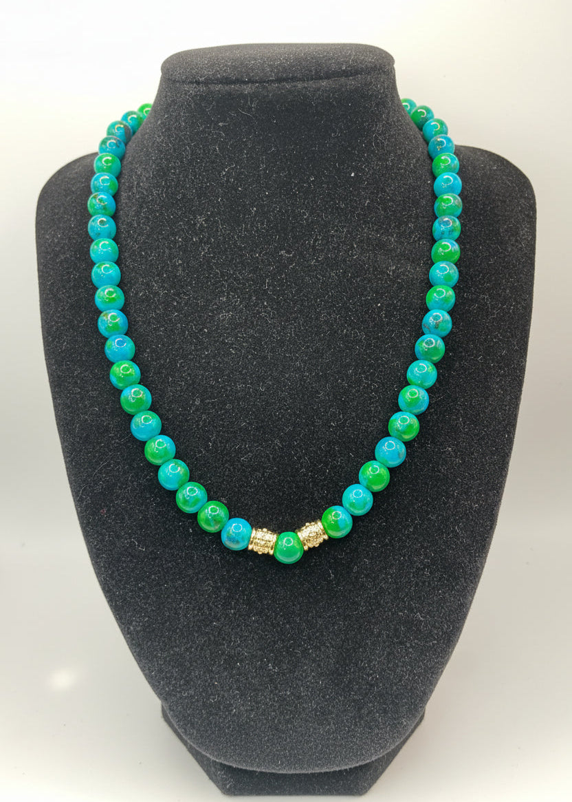 A beaded necklace with alternating bright blue and green round beads, accented by two small gold-toned spacer beads, displayed on a black velvet jewelry bust.