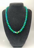 A beaded necklace with alternating bright blue and green round beads, accented by two small gold-toned spacer beads, displayed on a black velvet jewelry bust.