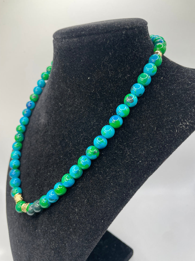 A beaded necklace with alternating turquoise blue and green marbled beads, accented by small gold spacers, displayed on a black velvet jewelry bust.