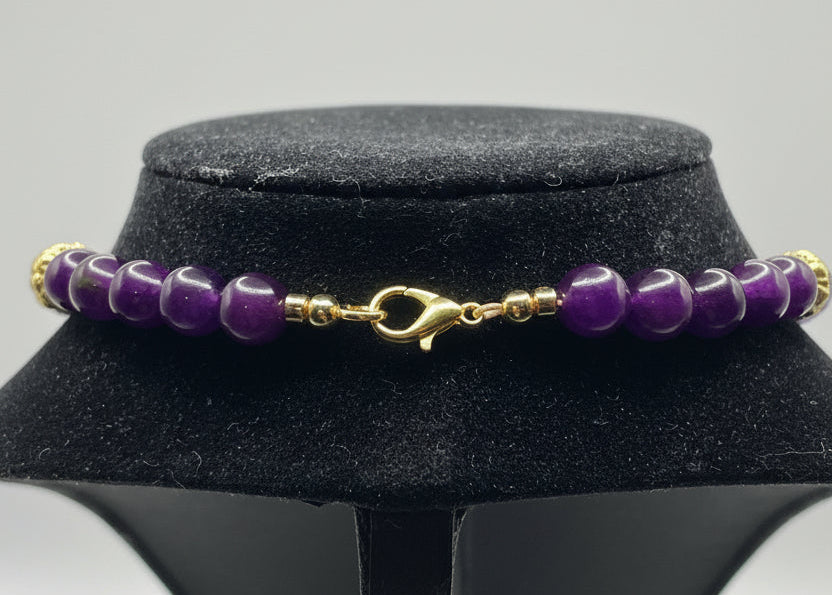 A beaded bracelet with polished purple beads and gold-toned spacer beads and clasp, displayed on a black velvet jewelry stand against a white background.