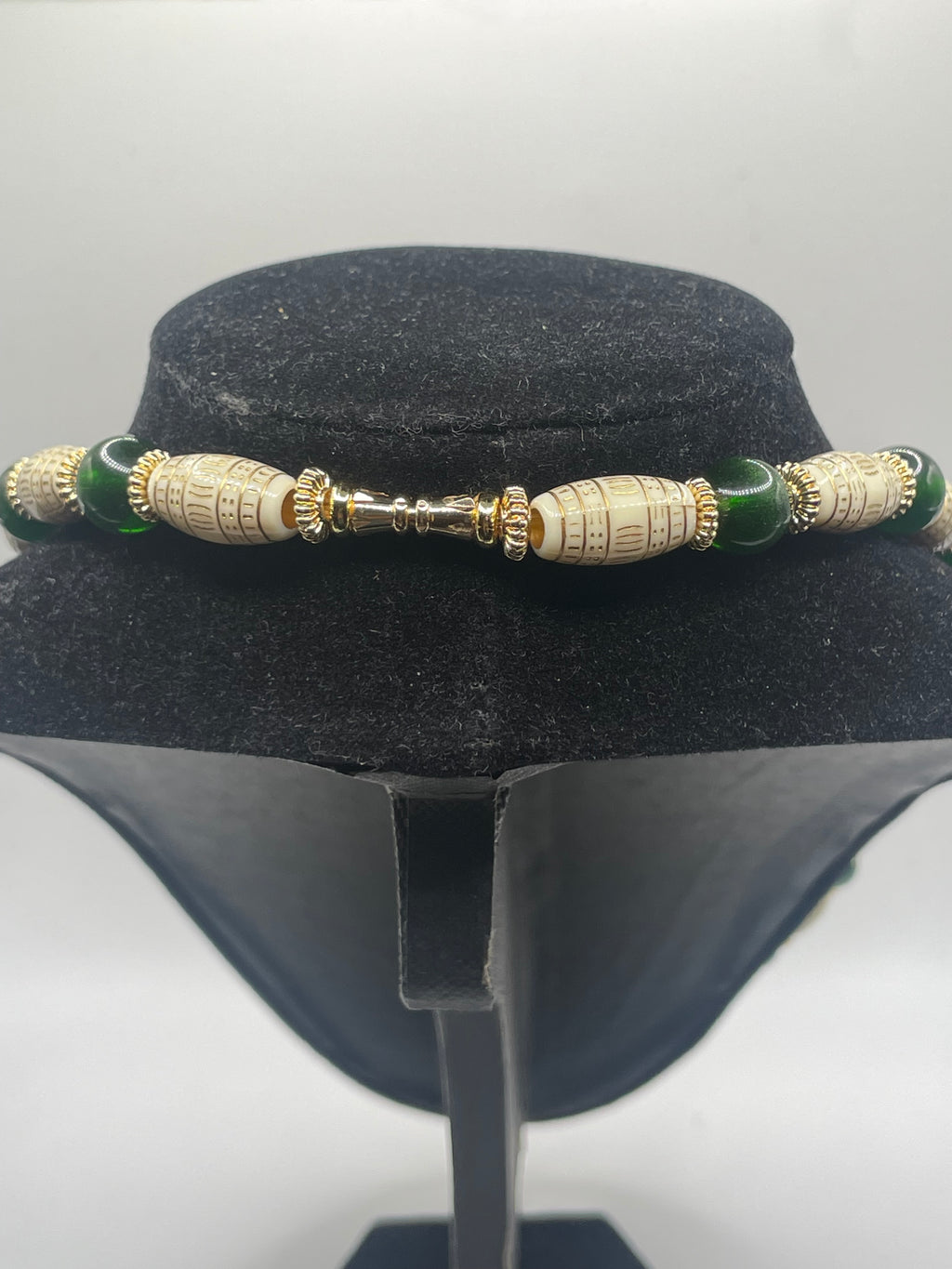 A beaded bracelet displayed on a black velvet jewelry bust. The bracelet features dark green round beads, large cream-colored cylindrical beads with gold-toned script details, and a central gold-toned clasp.