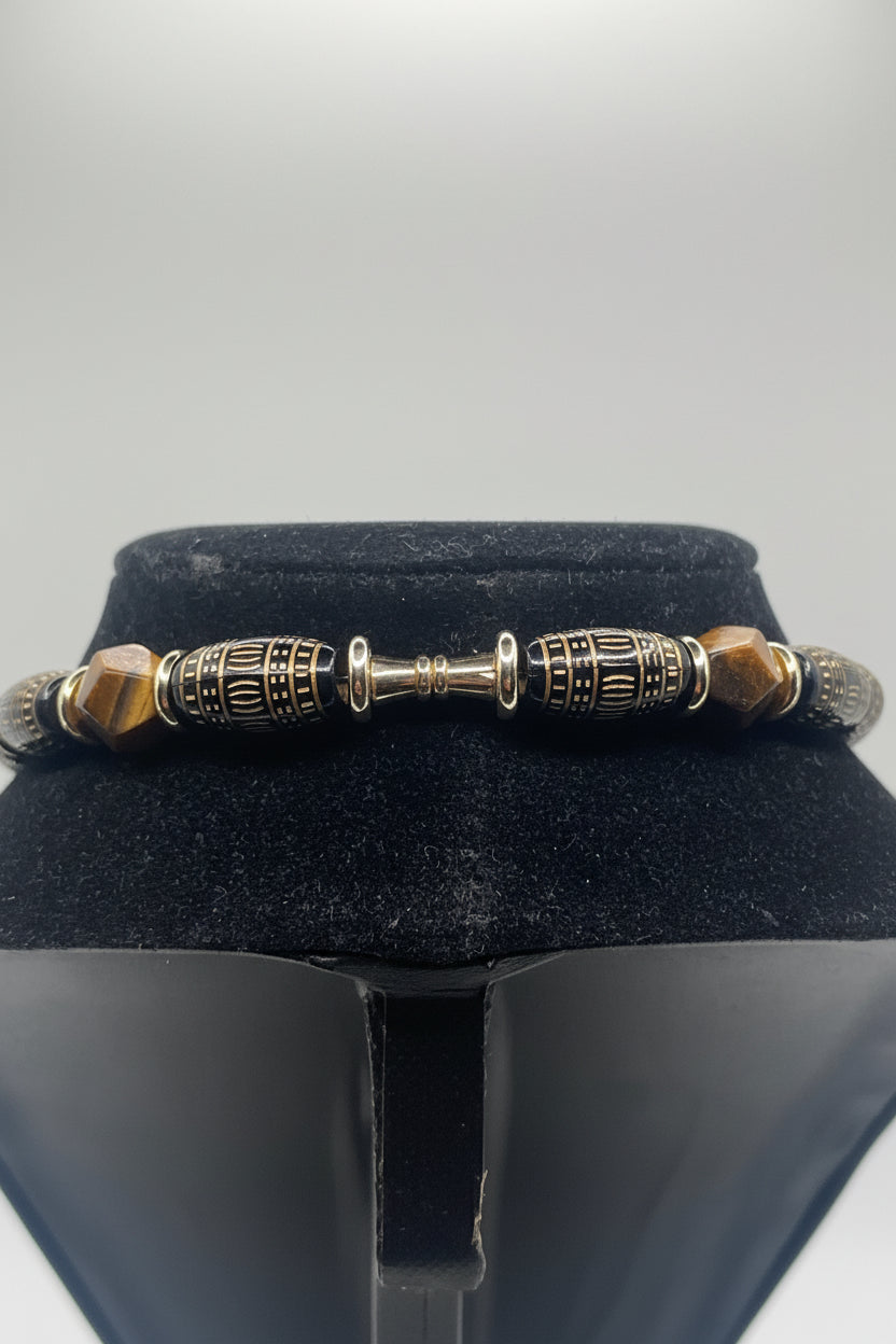 A beaded bracelet displayed on a black velvet jewelry bust. The bracelet features metallic gold-toned cylindrical beads with black and gold geometric patterns, a central gold-toned connector, and faceted brown beads on either side.