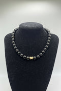 A beaded bracelet with dark gray to black polished round beads and a small gold-toned square charm, displayed on a black jewelry bust against a light gray background.