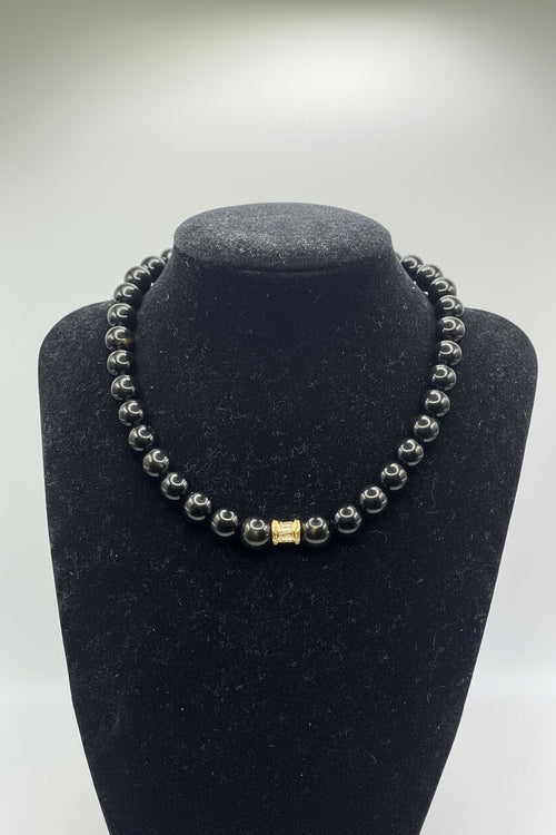A beaded bracelet with dark gray to black polished round beads and a small gold-toned square charm, displayed on a black jewelry bust against a light gray background.