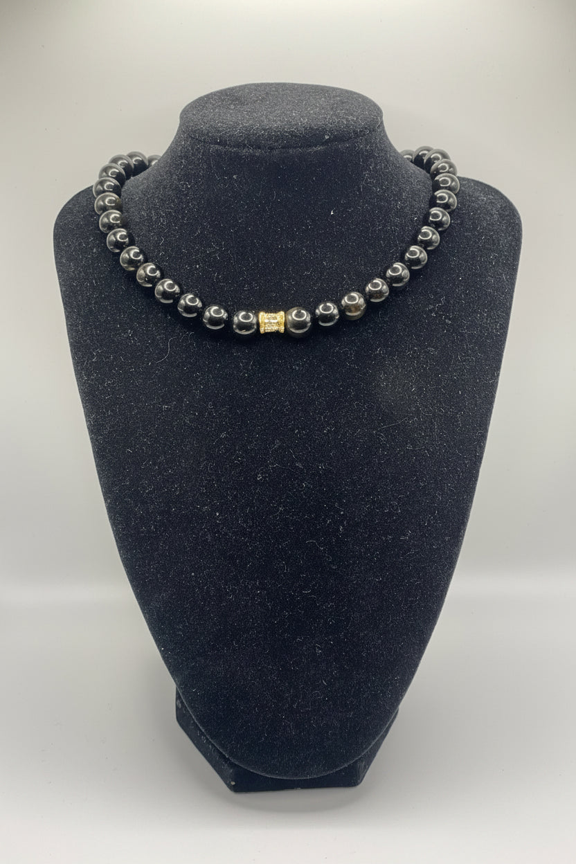 A beaded bracelet with dark gray to black round beads and a small gold rectangular accent bead, displayed on a black velvet jewelry bust against a light gray background.