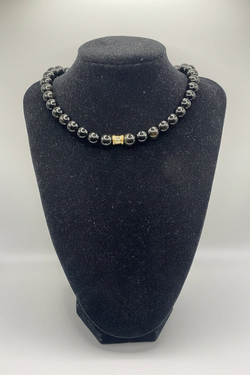 A beaded bracelet with dark gray to black round beads and a small gold rectangular accent bead, displayed on a black velvet jewelry bust against a light gray background.