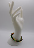 A beaded stretch bracelet with green, yellow, orange, and black beads, displayed on a white ceramic mannequin hand against a light gray background.