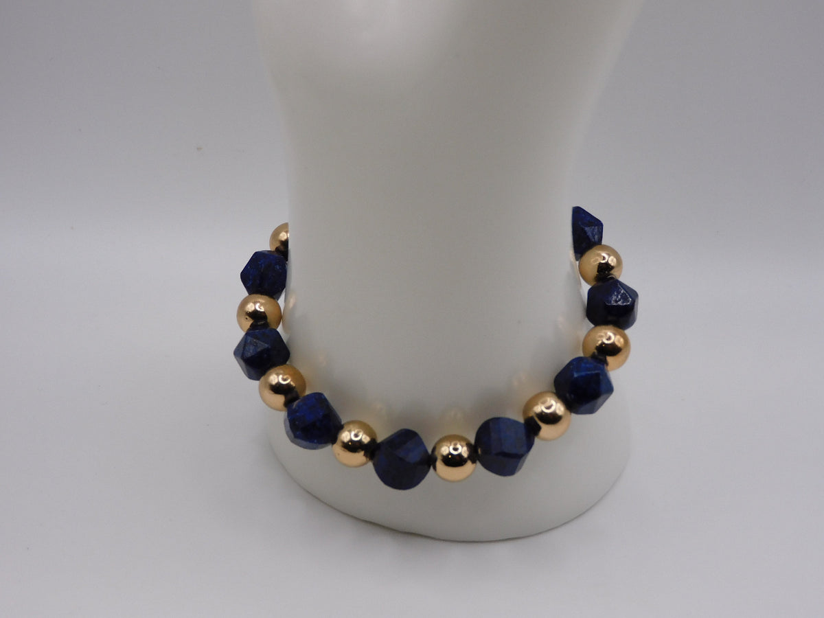 A beaded stretch bracelet featuring faceted dark blue beads and polished gold metallic beads, displayed on a white mannequin stand against a neutral light gray background.