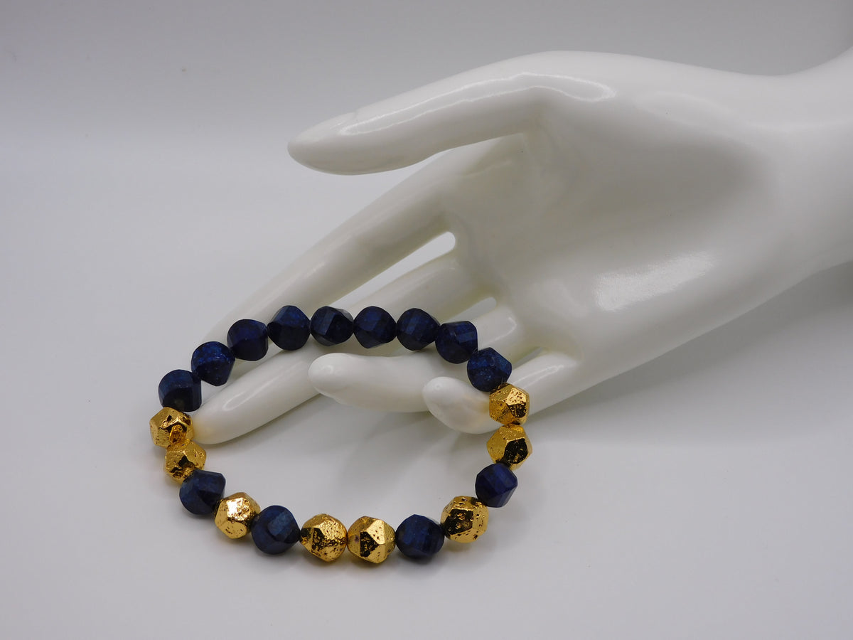 A beaded stretch bracelet with alternating dark blue faceted beads and textured gold beads, displayed on a white ceramic hand mannequin against a light gray background.