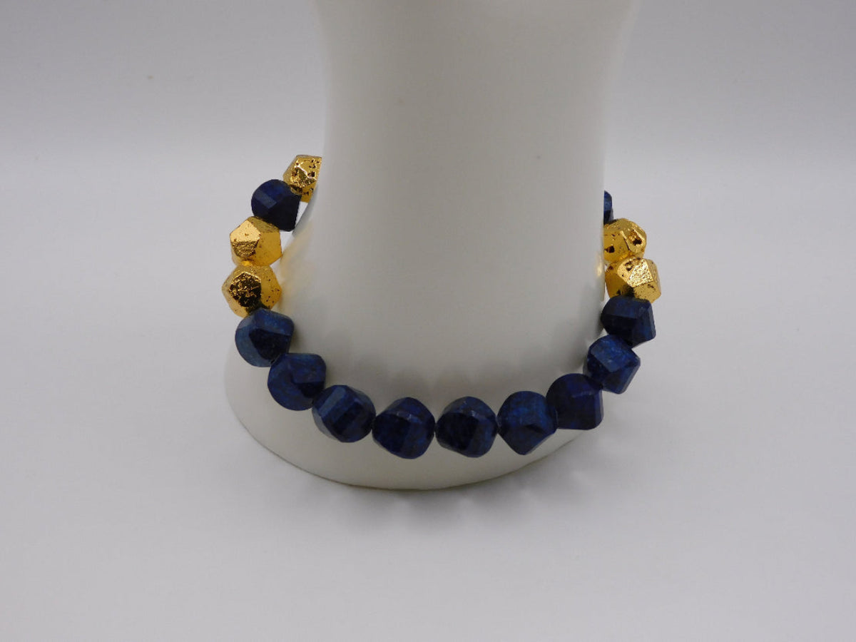 A beaded bracelet with faceted dark blue beads and textured gold spacer beads, displayed on a white cylindrical stand against a light gray background.
