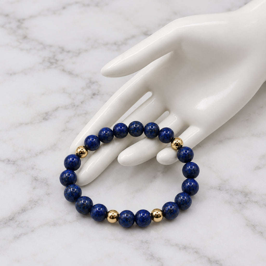 A beaded stretch bracelet made of dark blue lapis lazuli stones with small gold spacer beads, displayed on a white ceramic hand mannequin against a white background.