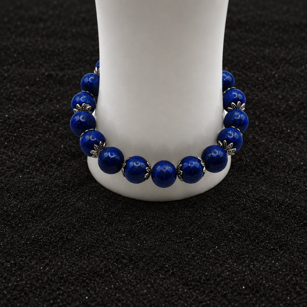 A beaded stretch bracelet made of dark blue lapis lazuli stones separated by small silver-toned metal spacers, displayed on a white cylindrical stand against a light gray background.