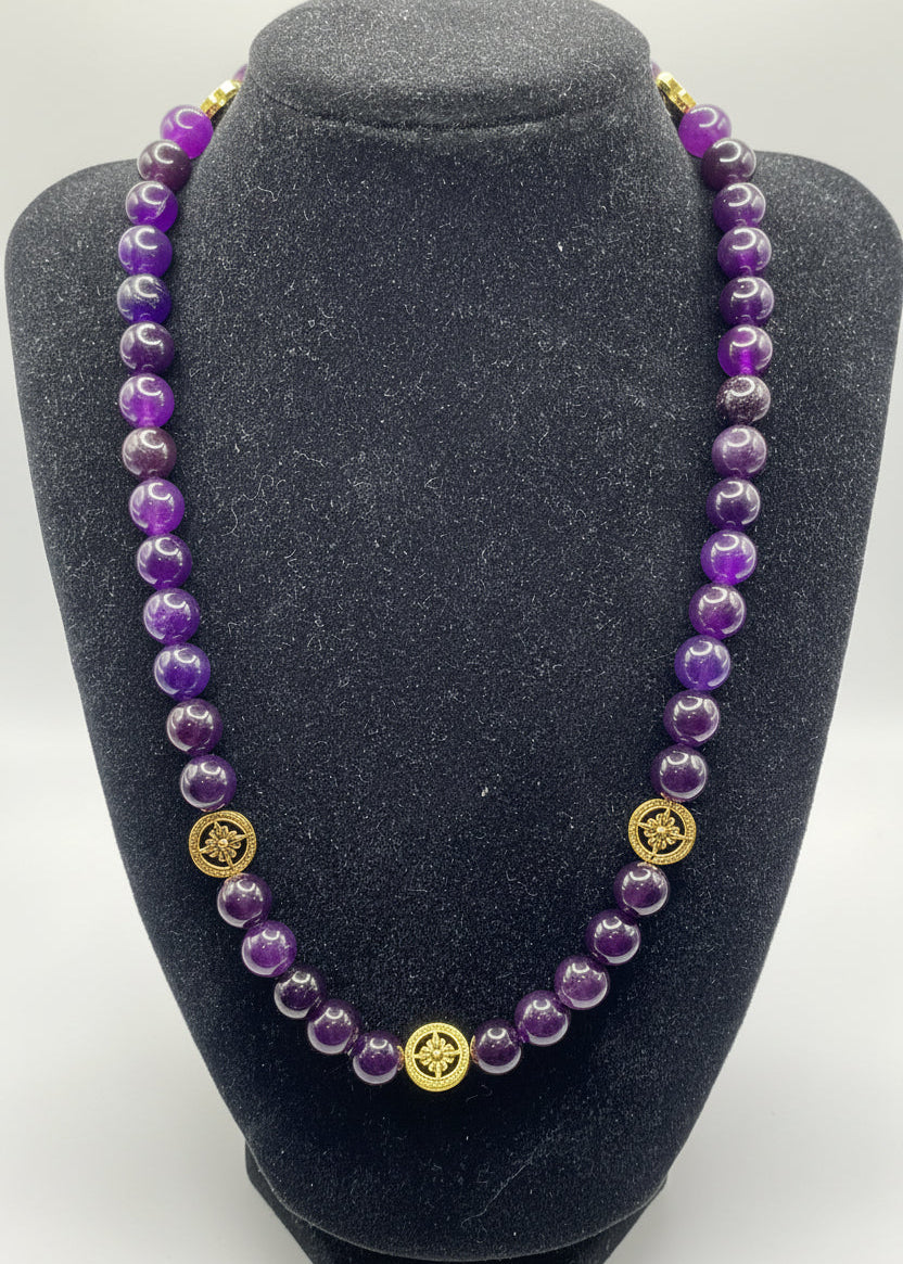 A beaded necklace made of polished purple amethyst beads with gold-toned circular spacer details, displayed on a black velvet jewelry bust against a light gray background.