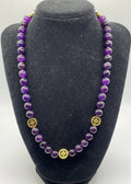 A beaded necklace made of polished purple amethyst beads with gold-toned circular spacer details, displayed on a black velvet jewelry bust against a light gray background.