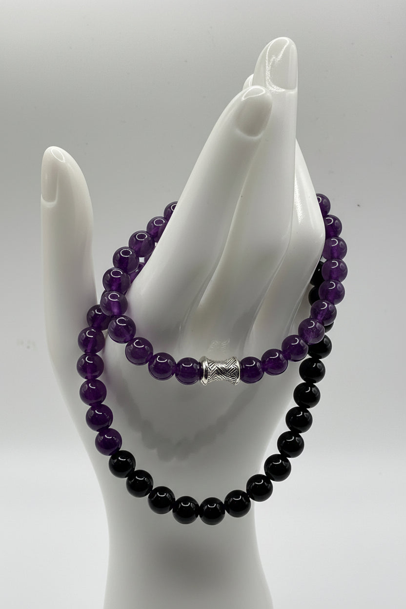 A beaded necklace displayed on a white mannequin hand, featuring alternating deep purple and glossy black beads with silver-toned cylindrical spacers.