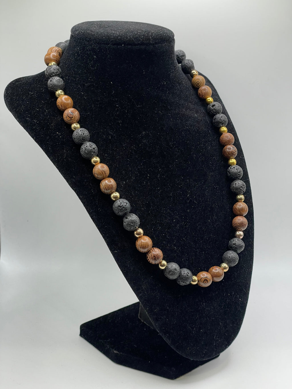 A beaded necklace made of alternating brown wood and dark gray lava stone beads, accented with small gold spacer beads, displayed on a black velvet jewelry bust.