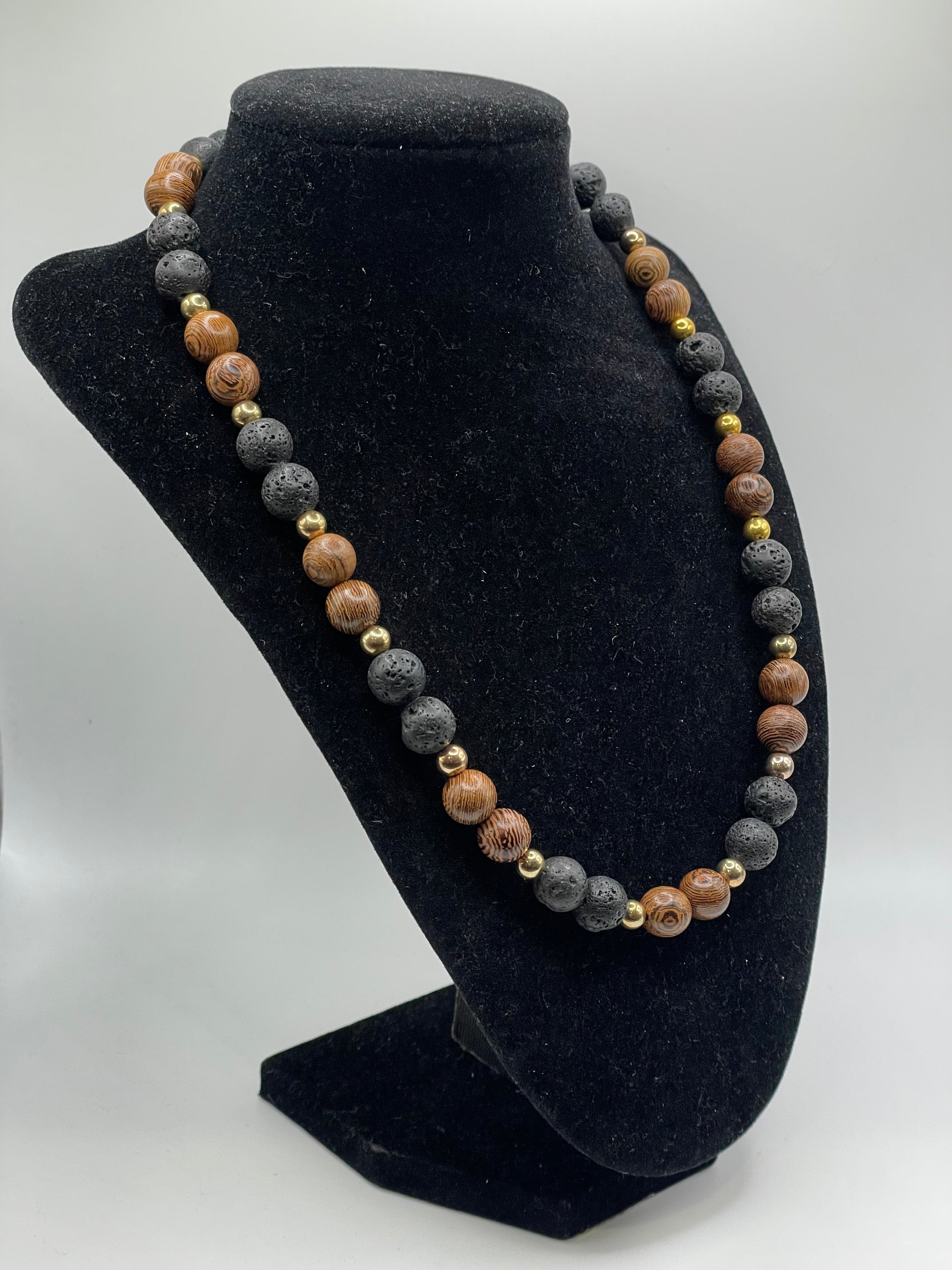 A beaded necklace made of alternating brown wood and dark gray lava stone beads, accented with small gold spacer beads, displayed on a black velvet jewelry bust.