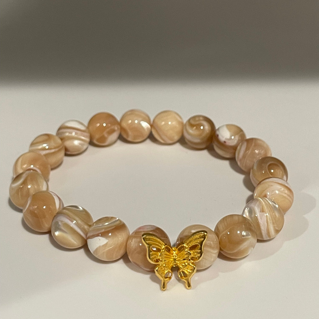 A beaded stretch bracelet made of glossy, marbled beige and white round beads, featuring a small gold butterfly charm, displayed against a neutral light gray background.