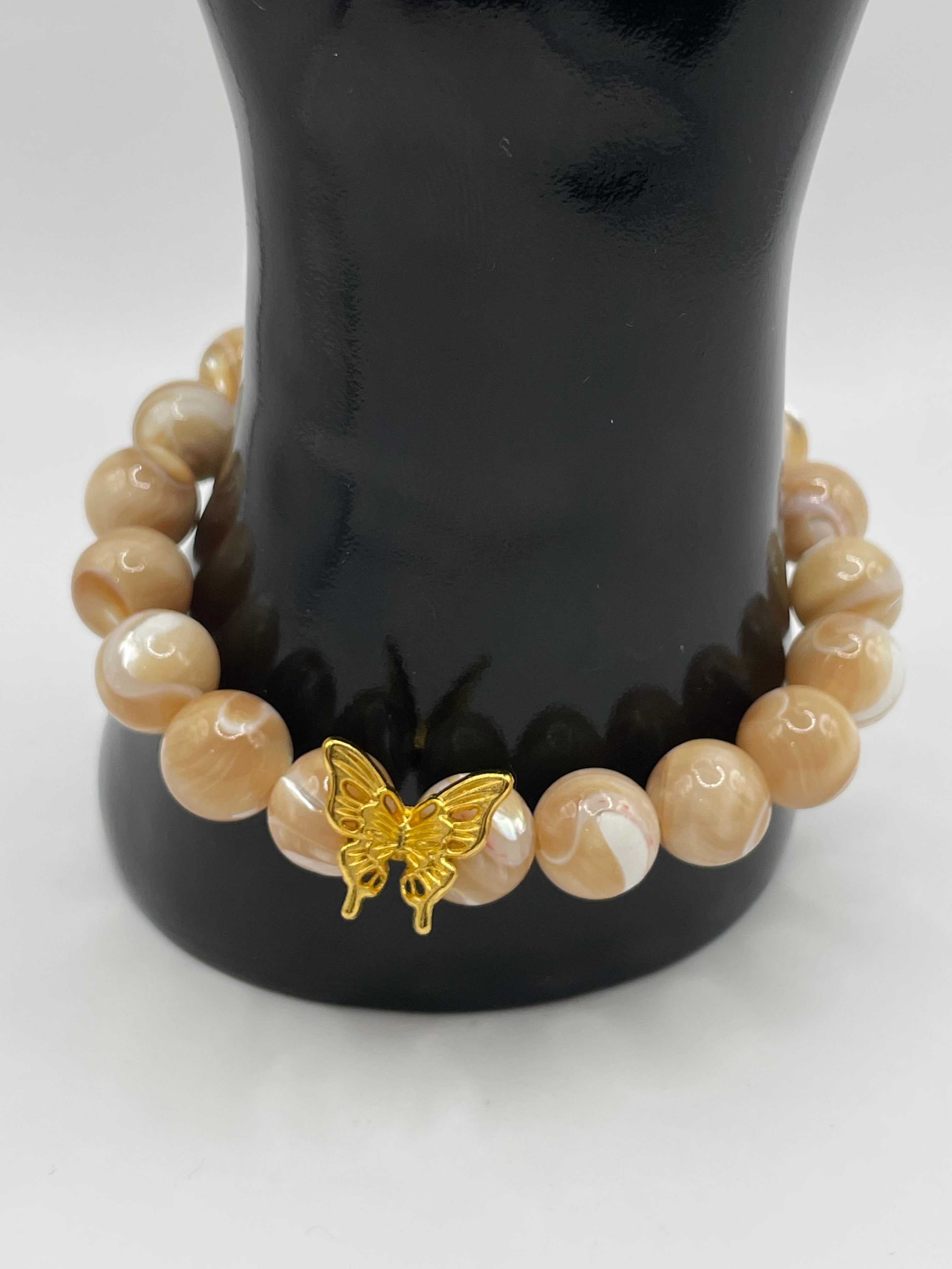 A beaded bracelet with smooth, round beige and cream marbled beads and a small gold butterfly charm, displayed on a black mannequin arm against a white background.