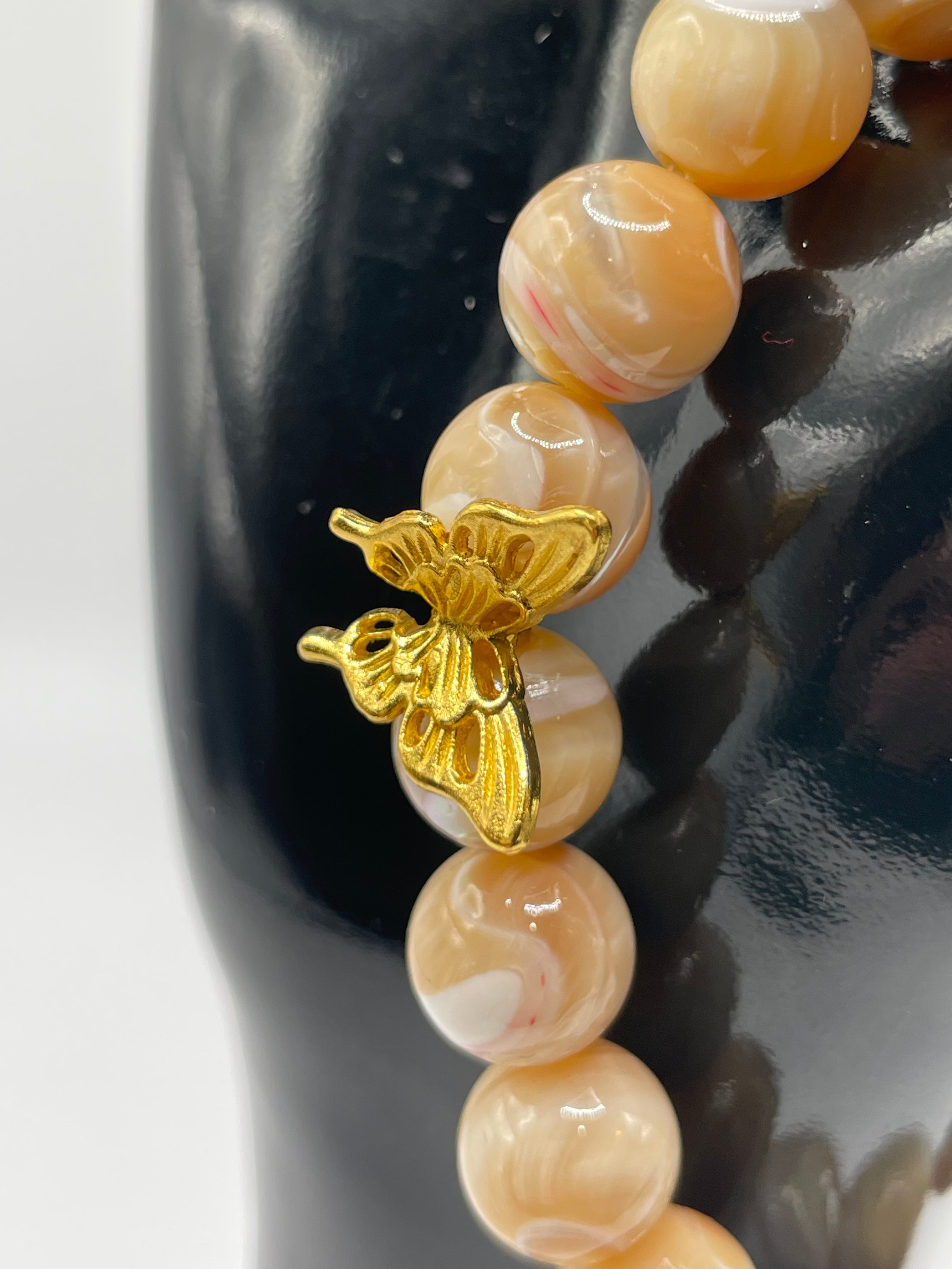 A close-up of a beaded bracelet with glossy peach and cream marbled beads and a small gold butterfly charm, displayed on a black surface.