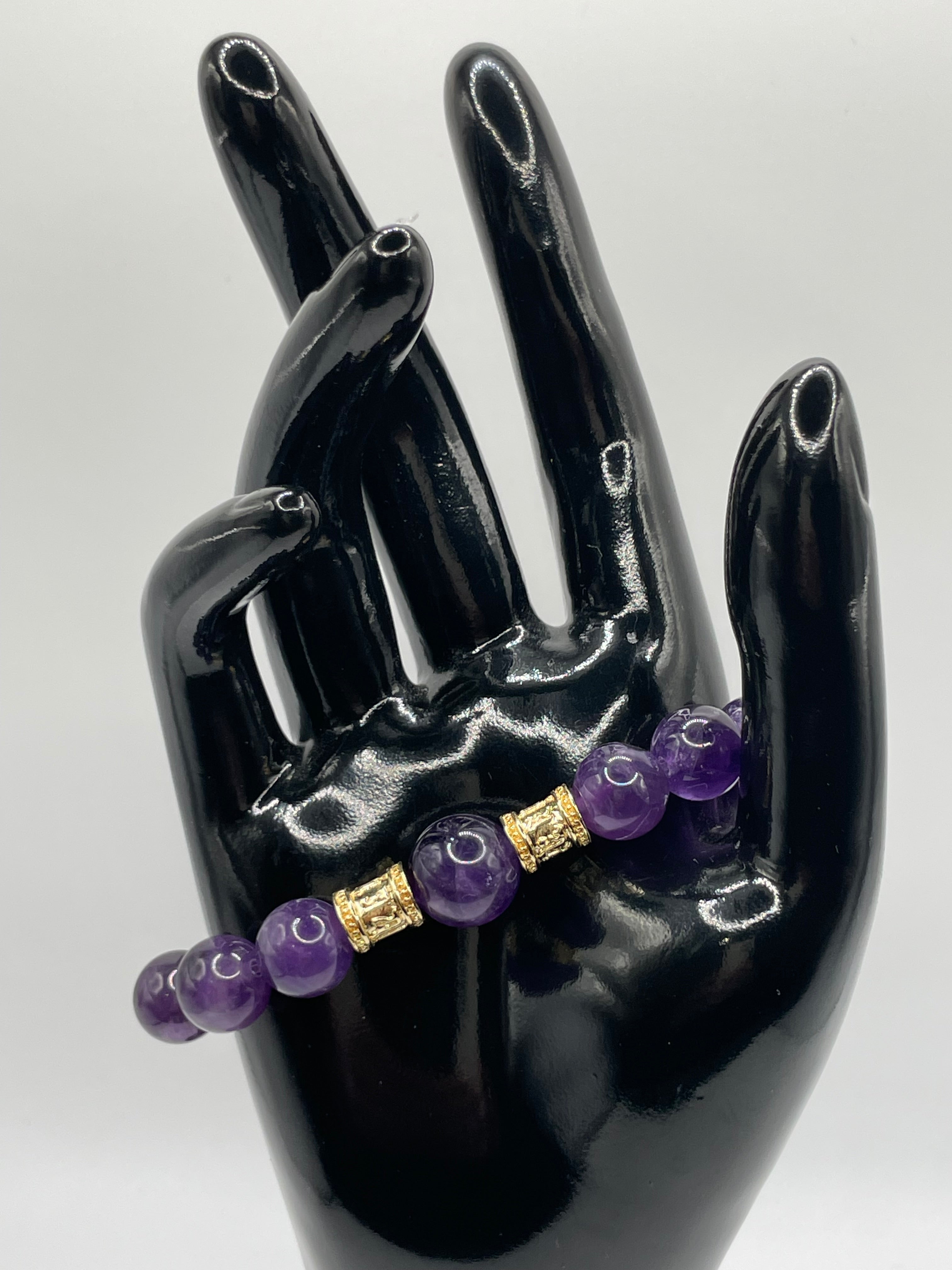 Black hand holding a bracelet with purple beads and gold accents on a white background