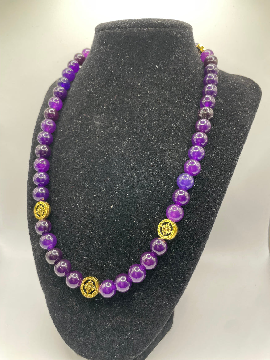 A beaded necklace made of purple and clear marbled beads with three gold circular charms, displayed on a black velvet jewelry bust against a white background.