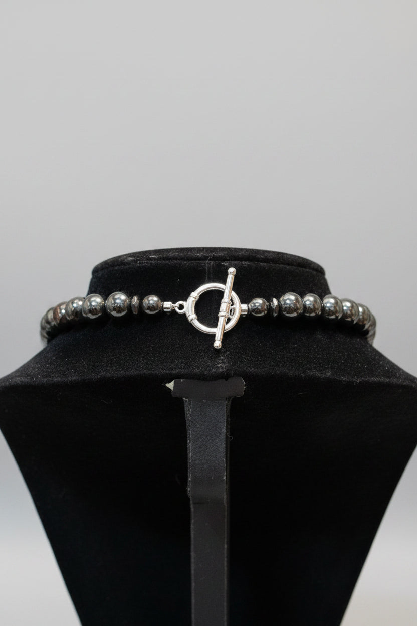 A beaded necklace back display with dark metallic beads and a silver toggle clasp, displayed on a black velvet jewelry bust.