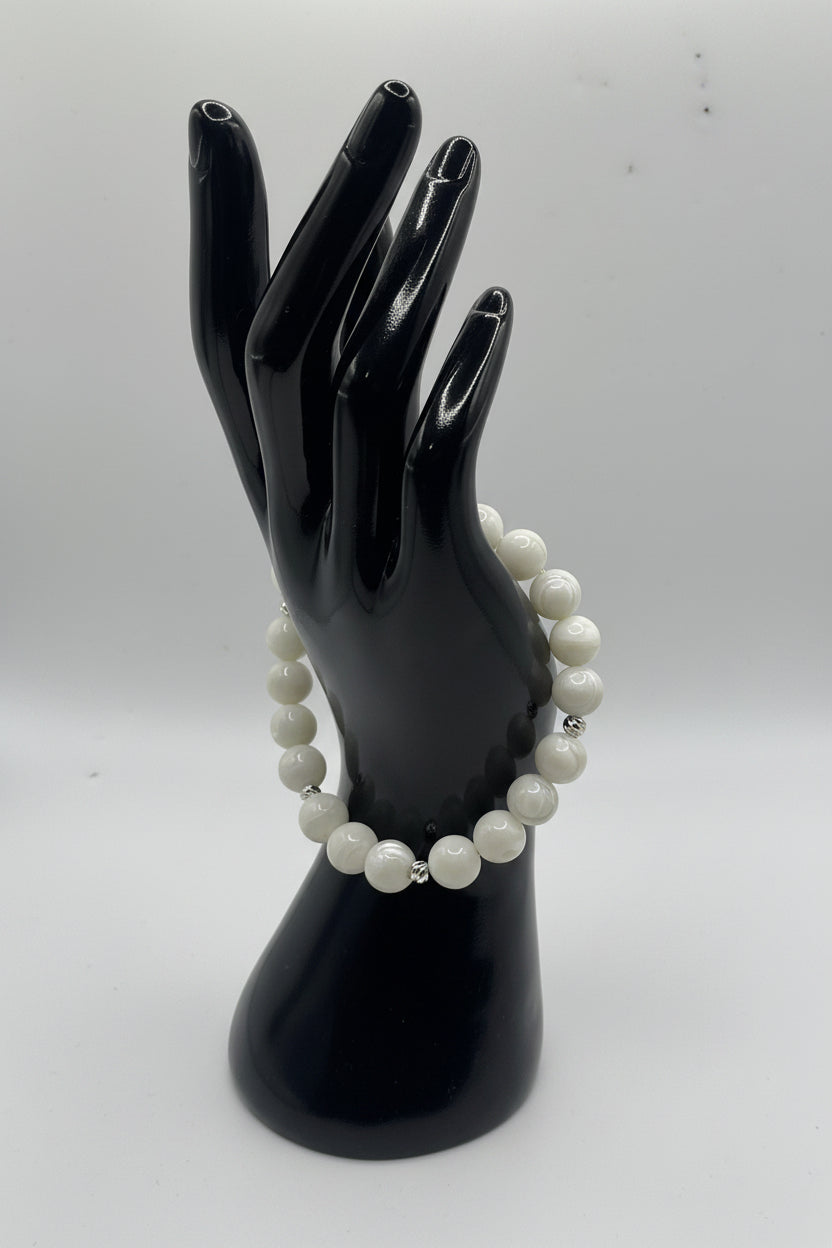 A beaded bracelet with white pearl-like round beads and small silver spacer beads, displayed on a glossy black hand-shaped mannequin against a light gray background.