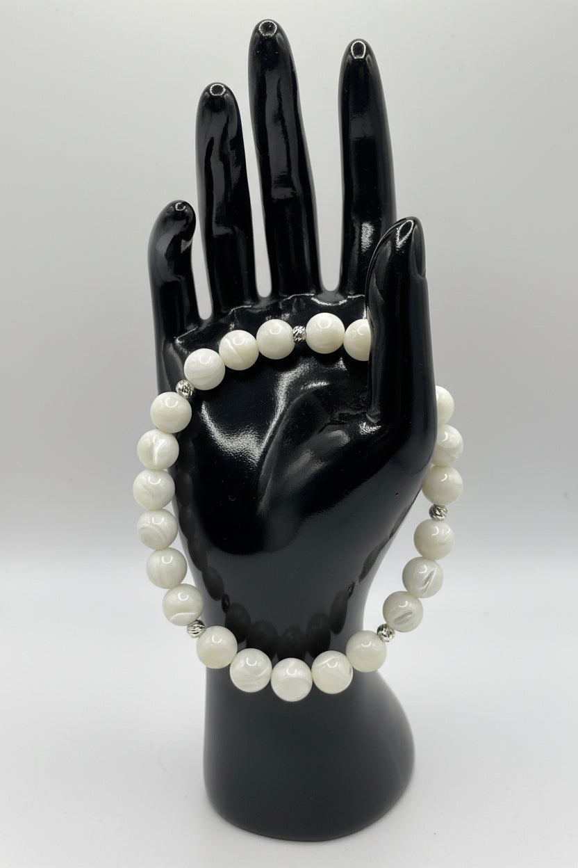 A beaded bracelet made of round white pearl-like beads separated by small silver spacer beads, displayed on a glossy black mannequin hand against a light gray background.