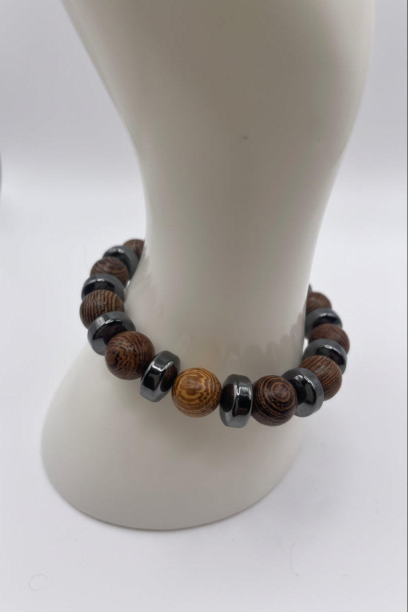 A beaded bracelet displayed on a white mannequin bust. The bracelet features alternating dark gray metallic beads, brown wood-grain beads, and one round light brown bead with darker concentric rings.