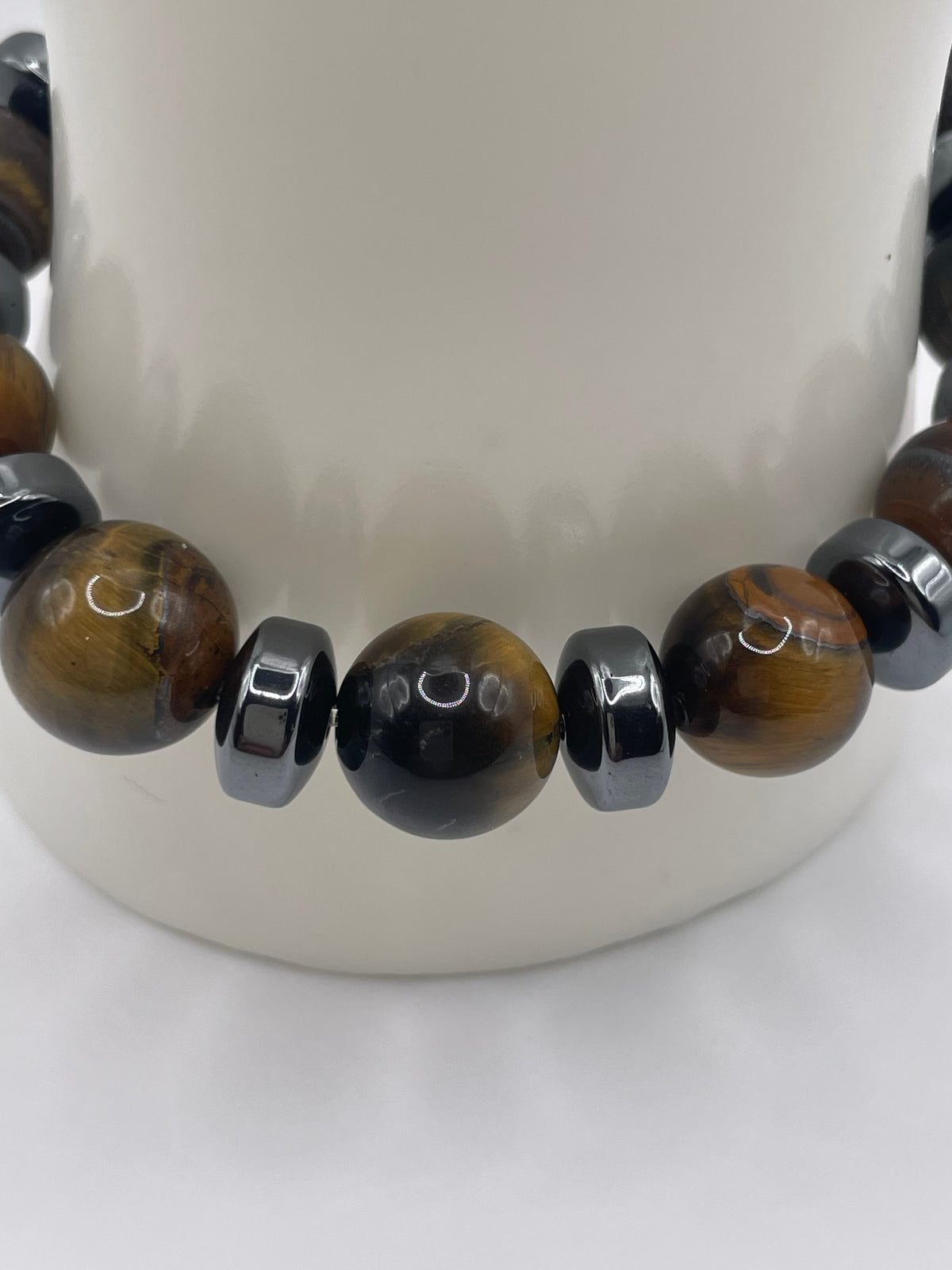 A close-up of a beaded necklace or bracelet featuring polished brown and black tiger's eye beads separated by shiny dark metallic spacer beads, displayed on a white curved stand.