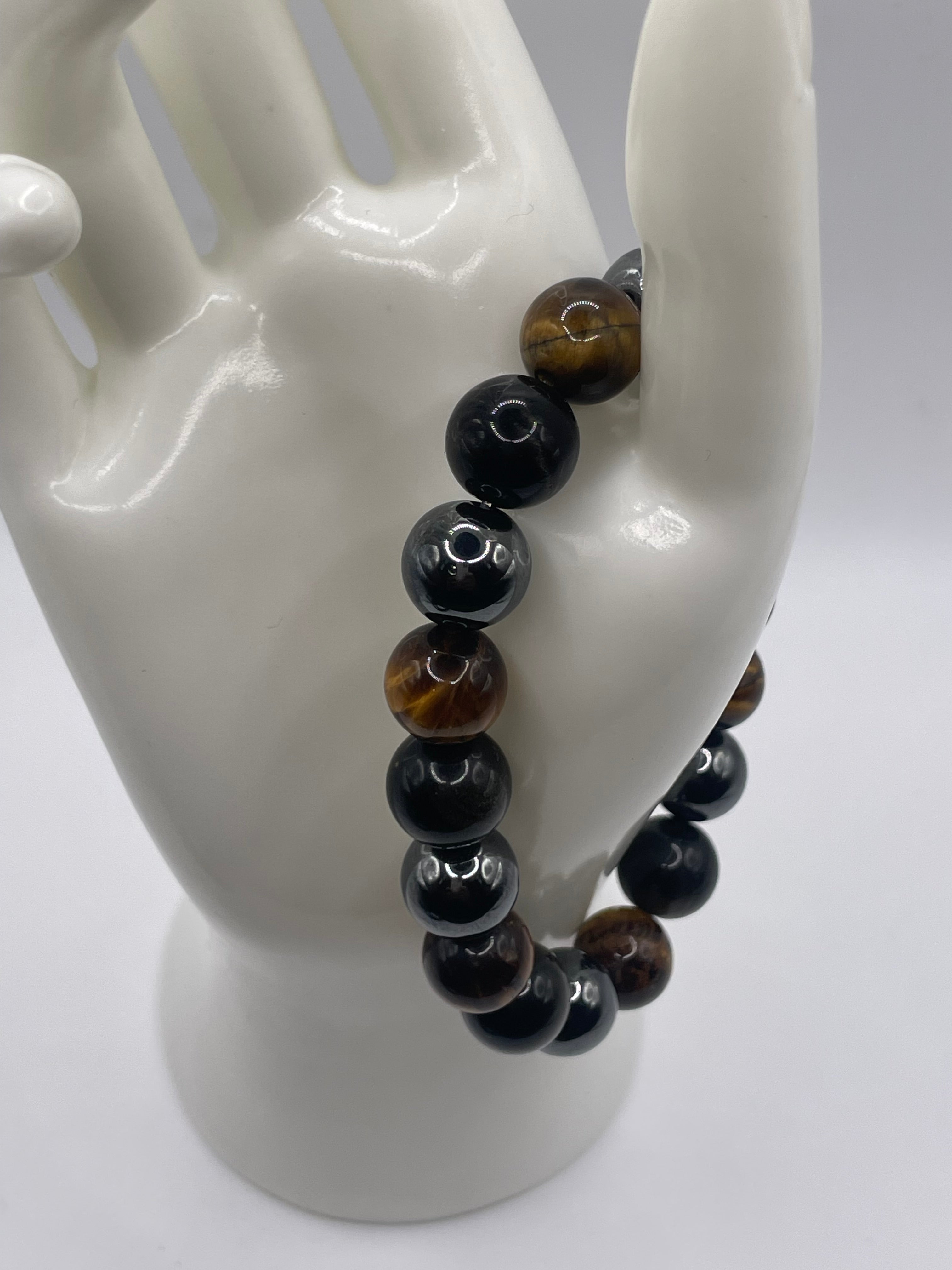 A beaded bracelet displayed on a glossy white ceramic hand-shaped stand. The bracelet features alternating glossy black round beads and tiger's eye beads showing brown and golden-brown striations.
