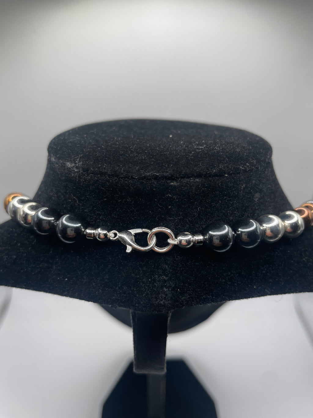 A beaded bracelet with alternating polished silver and dark gray beads, featuring a silver lobster clasp, displayed on a black velvet jewelry bust.