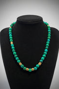 A beaded necklace made of turquoise and green marbled beads with two textured gold spacer beads, displayed on a black velvet jewelry bust.
