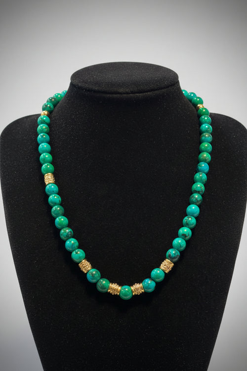 A beaded necklace made of turquoise and green marbled beads with two textured gold spacer beads, displayed on a black velvet jewelry bust.