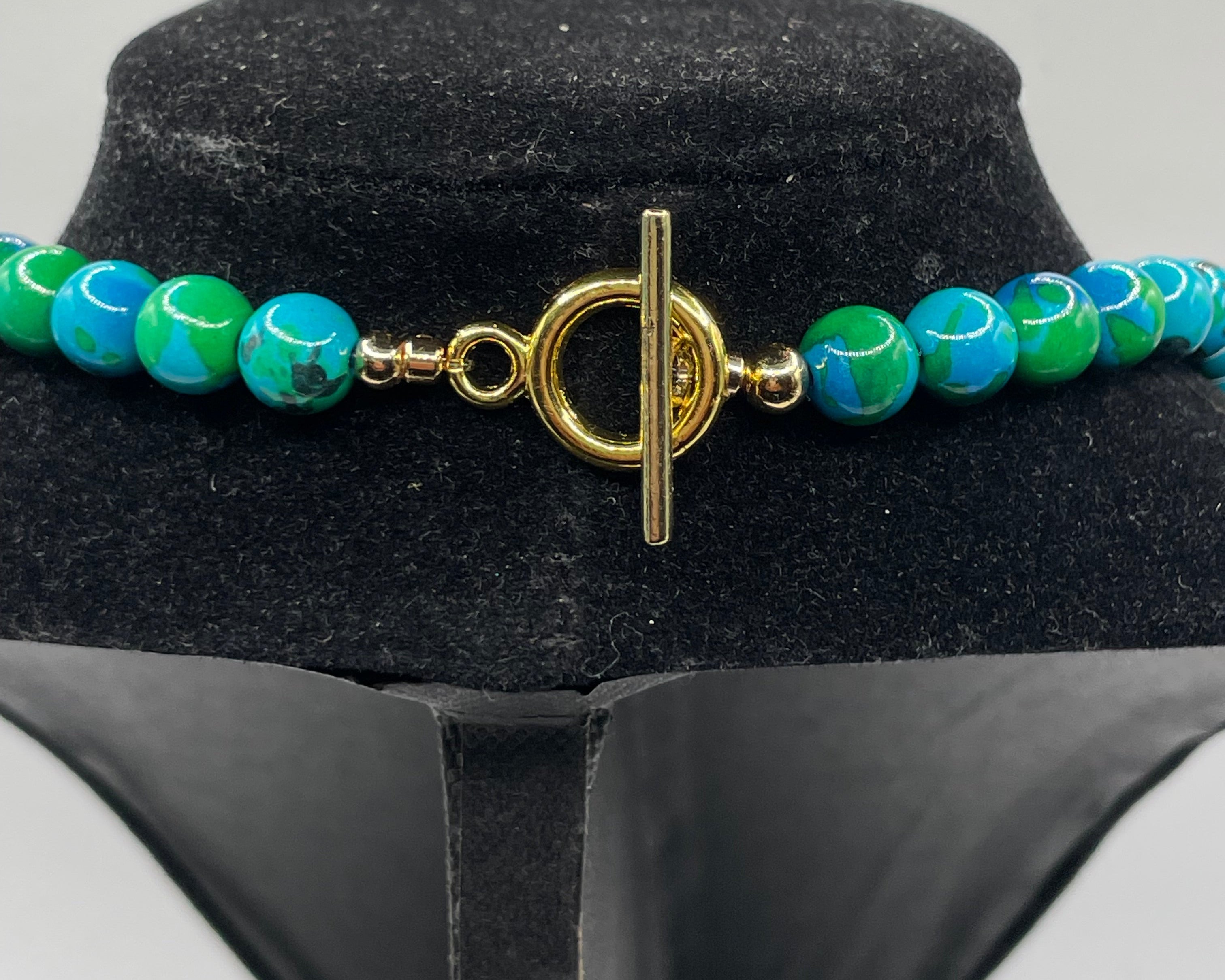 A beaded necklace with turquoise and green marbled beads and a gold toggle clasp, displayed on a black velvet jewelry bust against a light gray background.