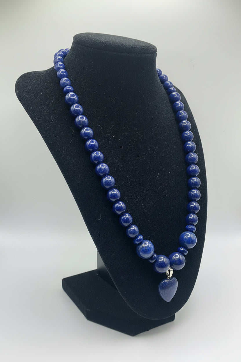 A beaded necklace made of polished blue lapis lazuli stones, featuring a heart-shaped pendant, displayed on a black velvet jewelry bust against a white background.