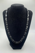 A beaded necklace displayed on a black velvet jewelry bust. The necklace features alternating matte black round beads and faceted dark gray square beads with silver-toned metal settings, photographed against a neutral light gray background.