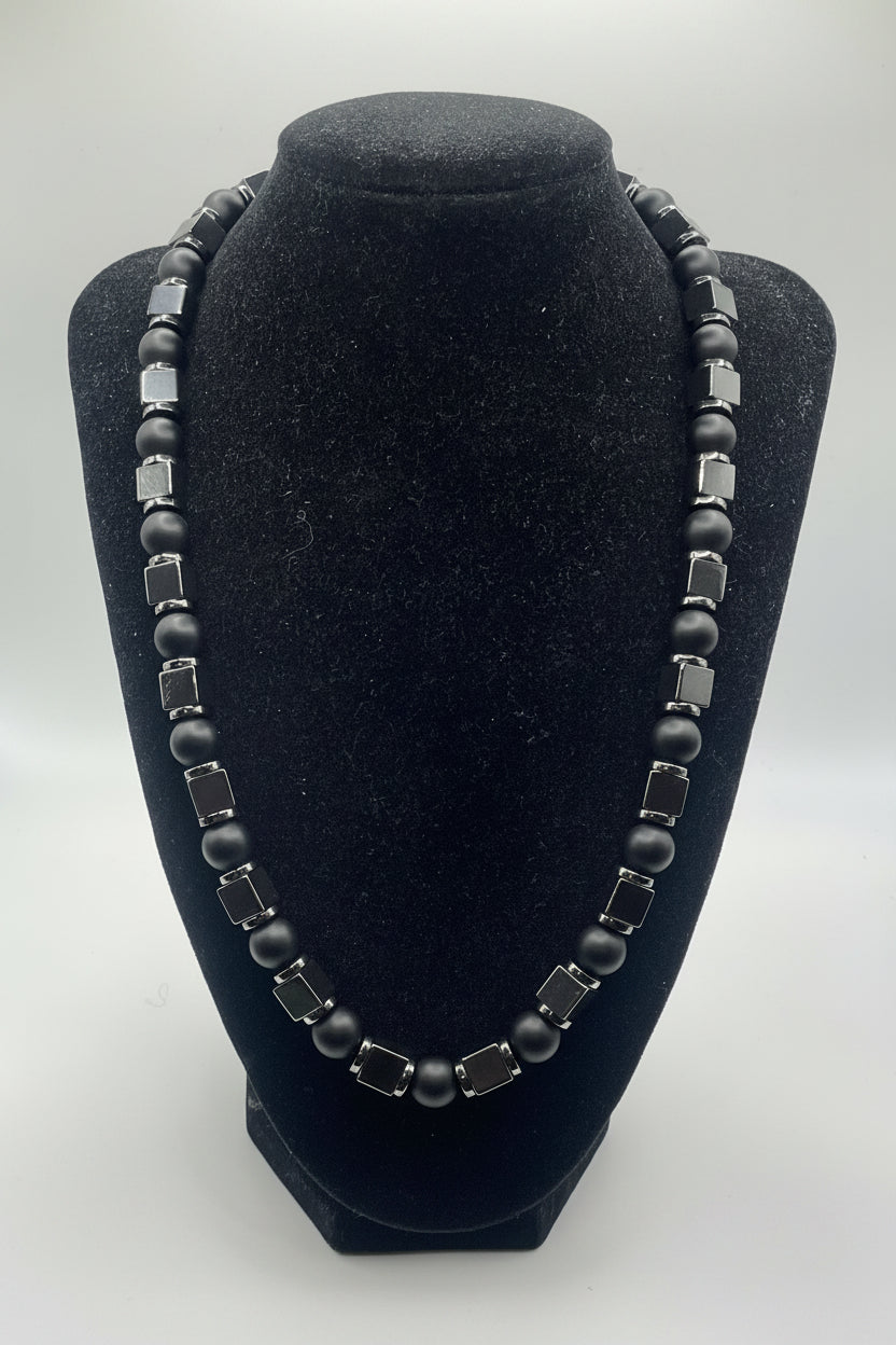 A beaded necklace displayed on a black velvet jewelry bust. The necklace features alternating matte black round beads and faceted dark gray square beads with silver-toned metal settings, photographed against a neutral light gray background.