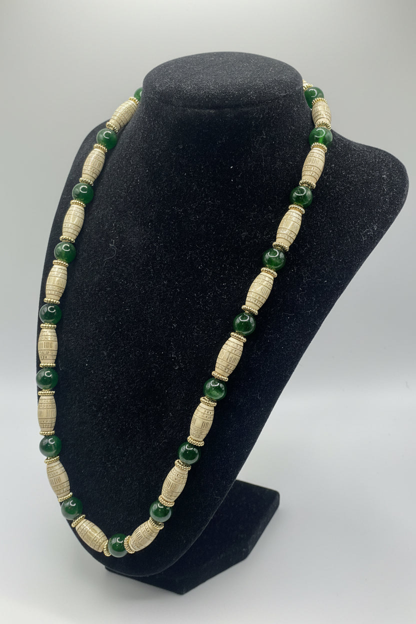A beaded necklace displayed on a black velvet jewelry bust. The necklace features alternating textured cream-colored cylindrical beads and glossy dark green round beads, separated by small gold-toned spacer rings.