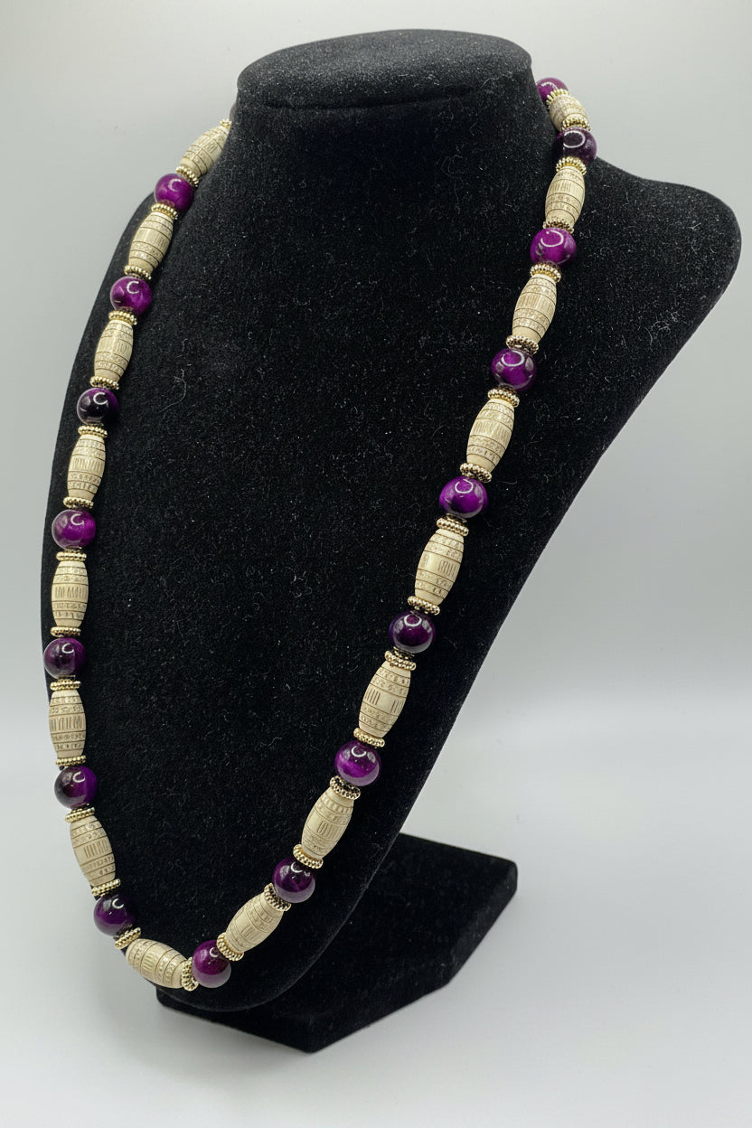 A beaded necklace displayed on a black velvet jewelry bust. The necklace features alternating light beige textured cylindrical beads and glossy purple round beads separated by small metallic gold spacer rings.