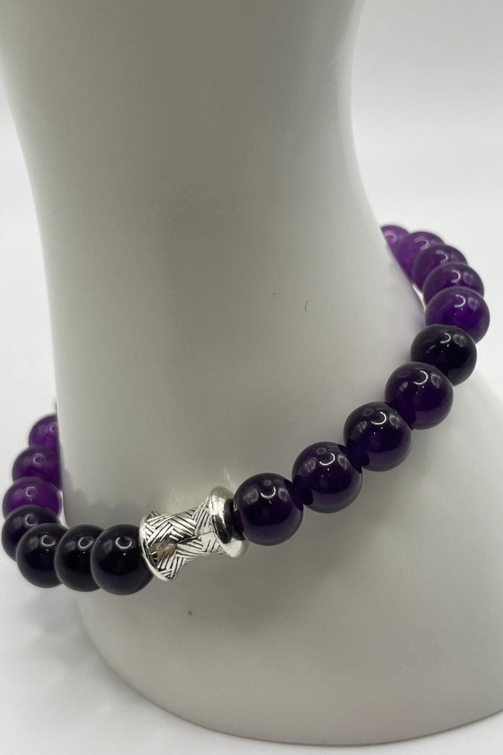 A beaded bracelet made of alternating glossy dark purple and black beads, featuring a textured silver spacer bead, displayed on a white mannequin stand.
