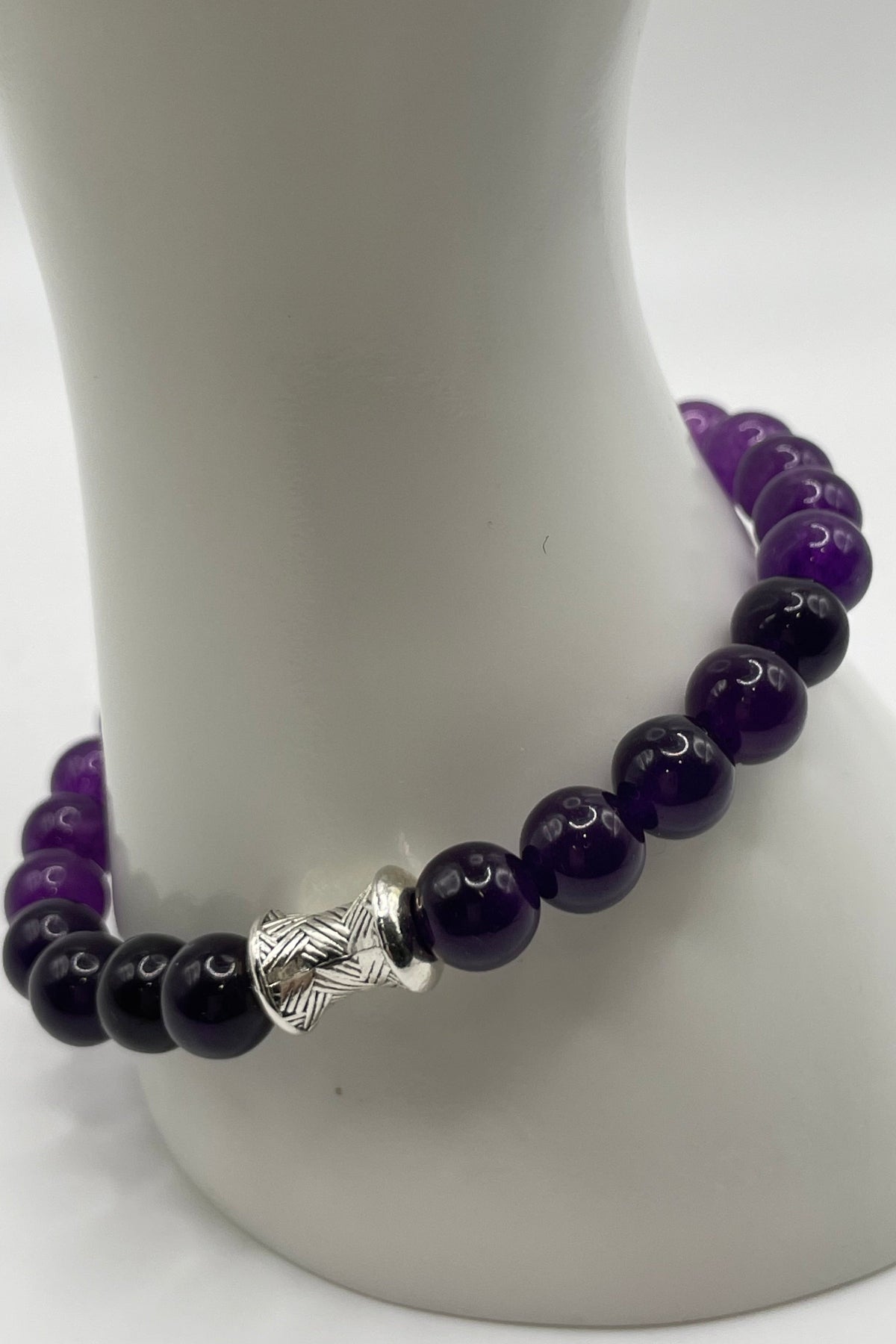 A beaded bracelet made of alternating glossy dark purple and black beads, featuring a textured silver spacer bead, displayed on a white mannequin stand.