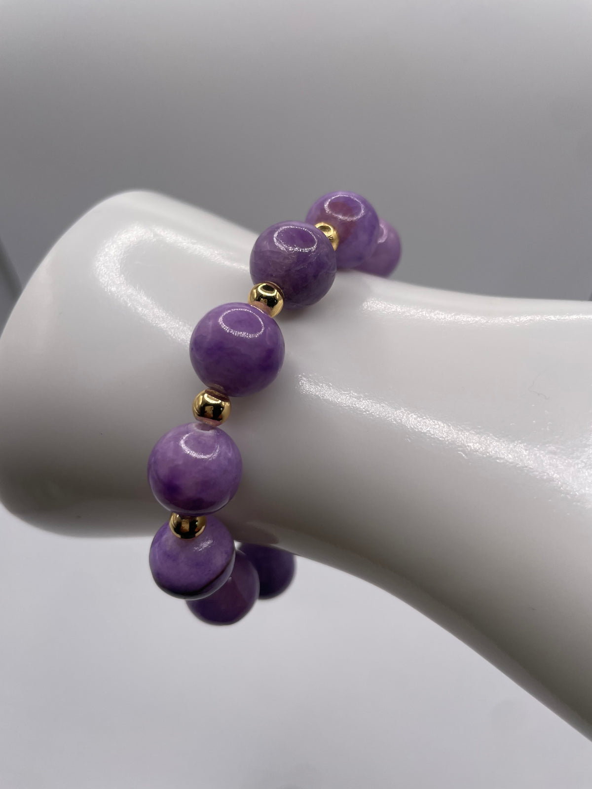 A beaded bracelet with polished purple round beads separated by small gold spacer beads, displayed on a white mannequin wrist against a soft gray background.