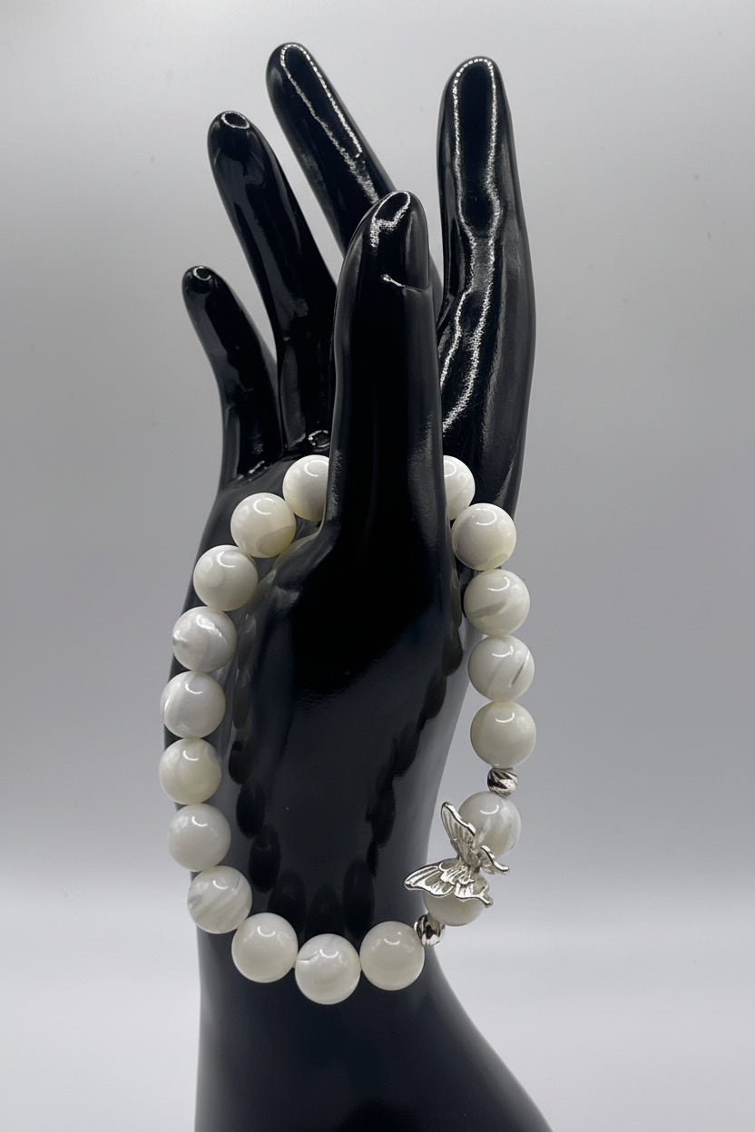 A beaded bracelet with white marbled round beads and a silver butterfly charm, displayed on a glossy black hand-shaped mannequin against a light gray background.