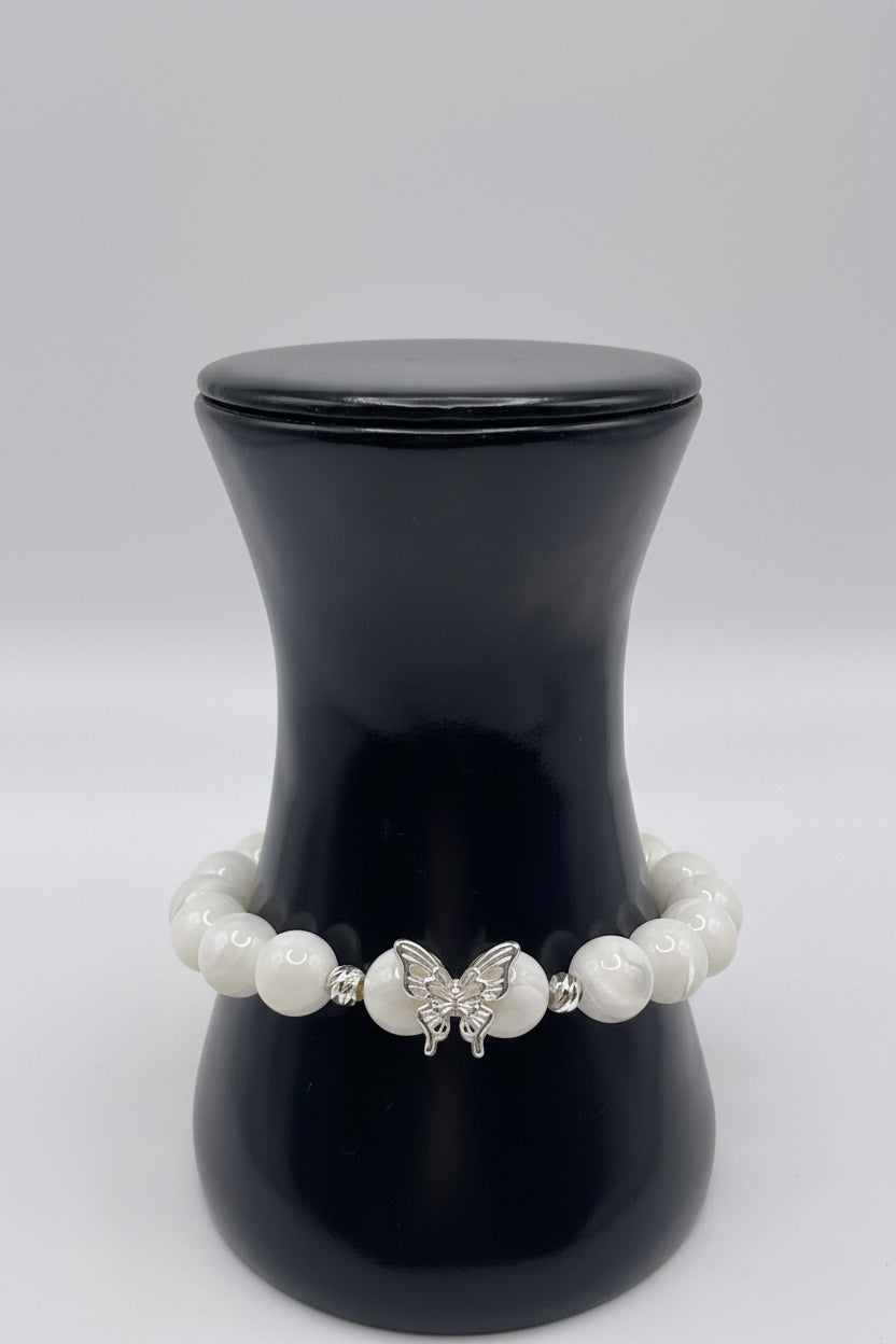 A beaded bracelet with white pearl-like round beads and a central silver butterfly charm, displayed on a black jewelry bust against a light gray background.