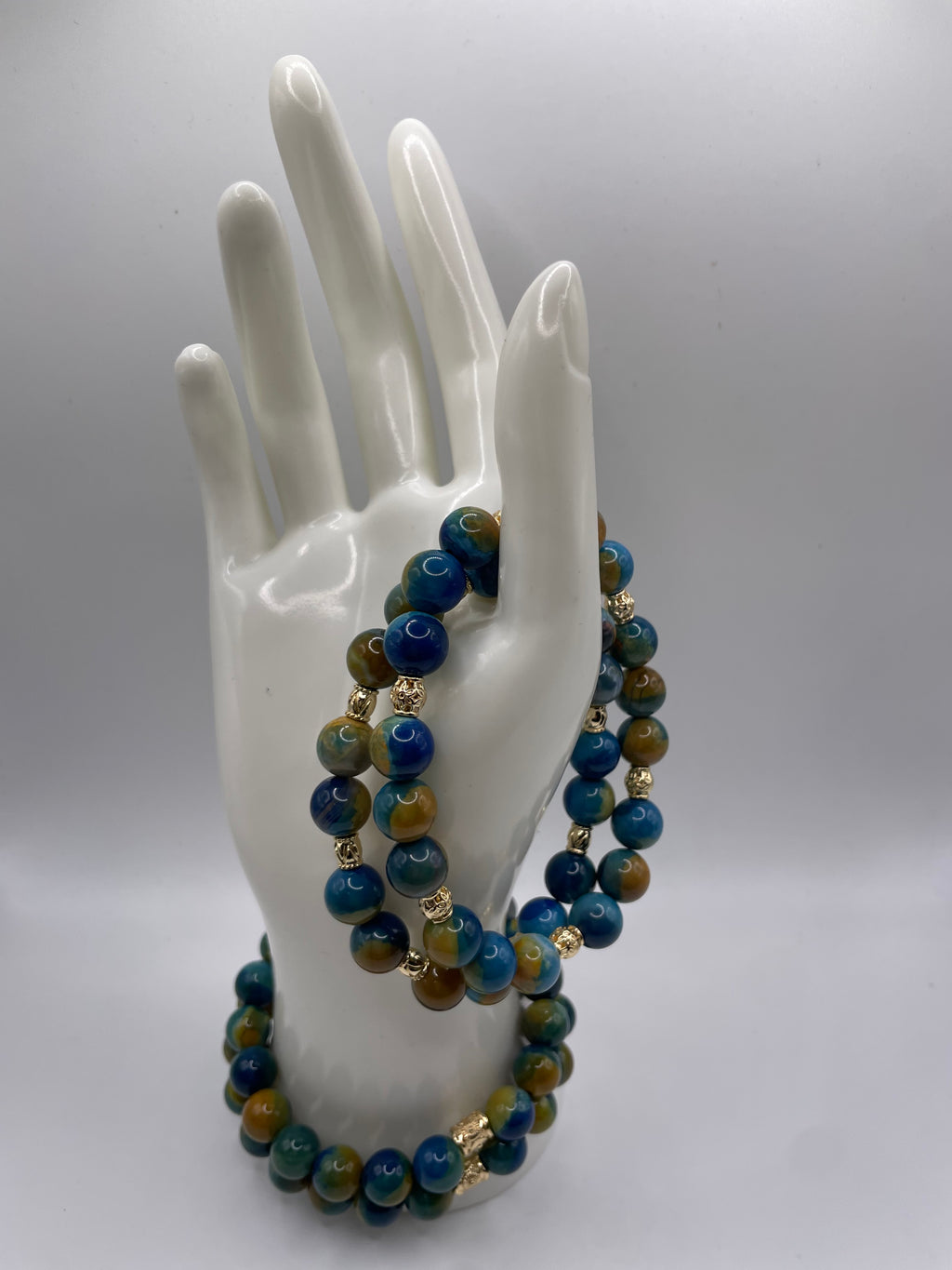 A beaded necklace and matching bracelet displayed on a white ceramic hand mannequin against a light gray studio background. The beads are primarily deep blue with green and brown marbled accents, separated by small gold spacer beads.
