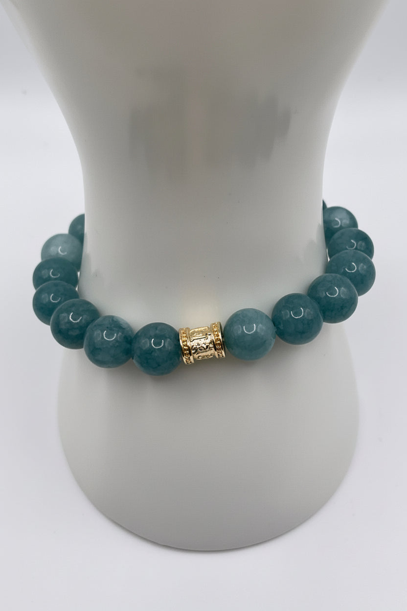 A beaded stretch bracelet with round teal-blue beads and a textured gold spacer, displayed on a white ceramic jewelry stand against a white background.