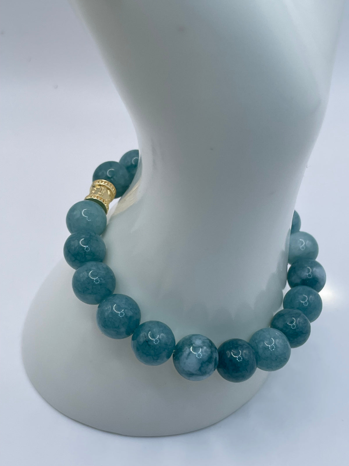 A beaded bracelet made of round teal-blue marbled beads with a small gold spacer bead, displayed on a white jewelry mannequin stand against a light gray background.